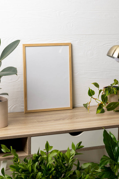 Vertical of empty wooden frame with copy space, lamp and plants in pots on table against white wall. Mock up frame template, interior design and decoration. - Photo, Image