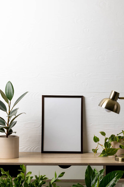 Vertical of black empty frame with copy space, lamp and plants in pots on table against white wall. Mock up frame template, interior design and decoration. - Photo, image