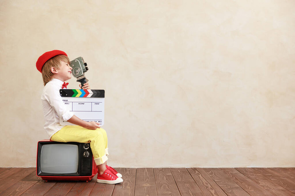 Funny boy holding vintage camera and clapper against grunge wall background. Kid sitting on old TV. Child playing at home - Photo, Image