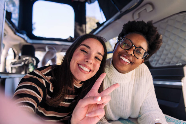 selfie photo of two happy young women having fun in a camper van, concept of weekend getaway with best friend and road trip adventure - Фото, изображение