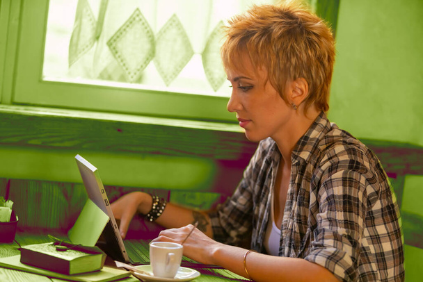 Mulher freelance com cabelo loiro curto e uma camisa xadrez funciona em um lugar público rústico com sua combinação laptop-tablet. Altamente focado e concentrado em escrever e pesquisar on-line como um profissional sério - Foto, Imagem
