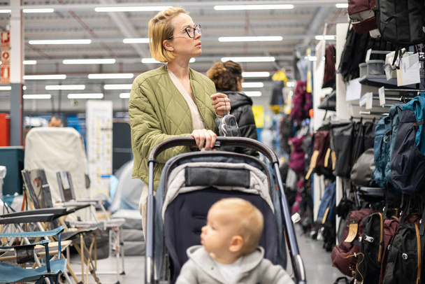 Casualy dressed mother choosing sporty shoes and clothes products in sports department of supermarket store with her infant baby boy child in stroller - Foto, Bild