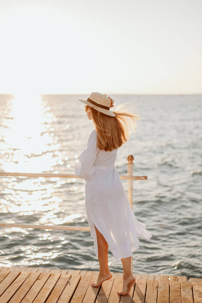 A young girl in a white dress and long hair, in sunglasses and a straw hat, stands on the pier against the backdrop of the ocean at sunset - Fotografie, Obrázek