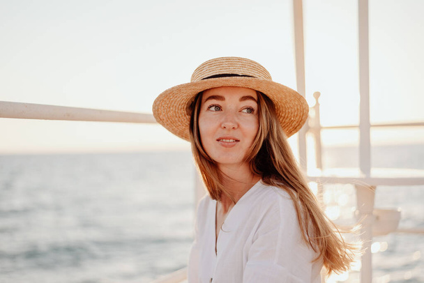 A beautiful young girl with long hair in a white dress and a straw hat smiles beautifully against the backdrop of the evening ocean. - Photo, Image