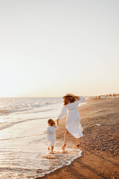 A little girl and a young mother in white dresses and a hat walk barefoot on the sand on the ocean at sunset. Wind blows hair - Photo, Image
