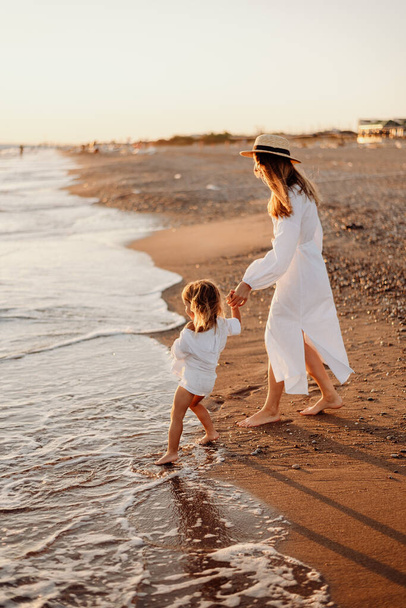 A little girl and a young mother in white dresses and a hat are walking on the water on the ocean at sunset. Deserted beach. - Photo, image