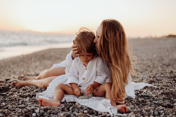 Uma jovem mãe e filha em vestidos brancos estão sentados em uma costa oceânica rochosa ao pôr-do-sol. Uma pequena filha olha para uma pedra em suas mãos, e sua mãe a beija - Foto, Imagem