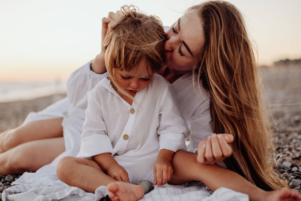 Une jeune mère et sa fille en robe blanche sont assises sur un rivage rocheux au coucher du soleil. Une petite fille regarde une pierre dans ses mains, et sa mère l'embrasse - Photo, image