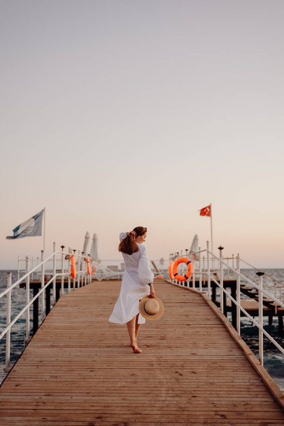 Nuori tyttö, jolla on pitkät hiukset valkoisessa mekossa, kävelee kohti merta puulaituria ja pitää hattua käsissään. Hiukset kehittää tuuli - Valokuva, kuva