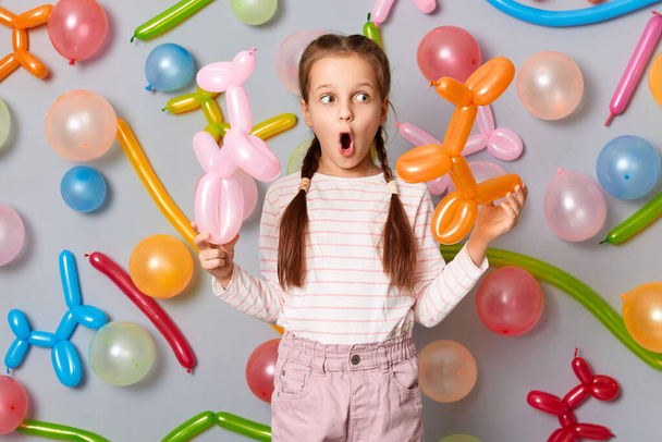 Funny shocked surprised little girl with pigtails standing against gray wall decorated with colorful balloons, looking away, sees surprise, excellent birthday present. - Zdjęcie, obraz