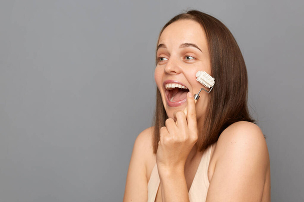 Amazed overjoyed young woman with brown hair massaging her face with jade rollers enjoying beauty skin care procedures posing isolated over gray background. - Valokuva, kuva