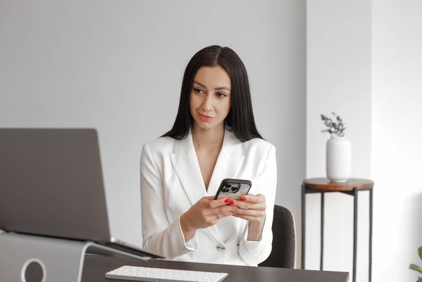 Front view of elegant caucasian woman with long hair sitting, browsing mobile phone and looking at laptop screen at home or at work. Indoors, Horizontal, copy-space - Foto, afbeelding