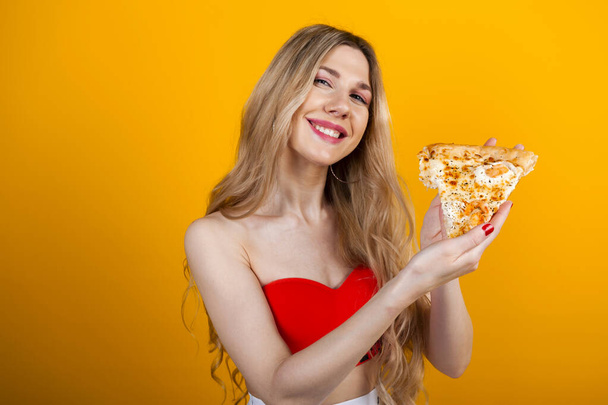 Ein schönes, fittes, junges blondes Mädchen posiert mit einer Pizzascheibe. Isoliert auf Gelb. - Foto, Bild