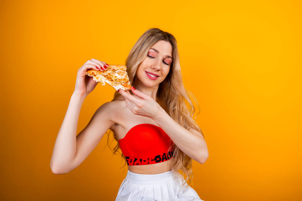 Красивая подтянутая молодая блондинка позирует с кусочком пиццы. Изолированный на желтый. - Фото, изображение
