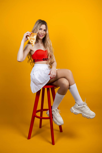 Ein wunderschönes, sinnlich sitzendes junges blondes Mädchen in kurzem Top und Rock sitzt auf einem Barhocker und posiert mit einem Pizzascheibchen. Isoliert auf Gelb. - Foto, Bild