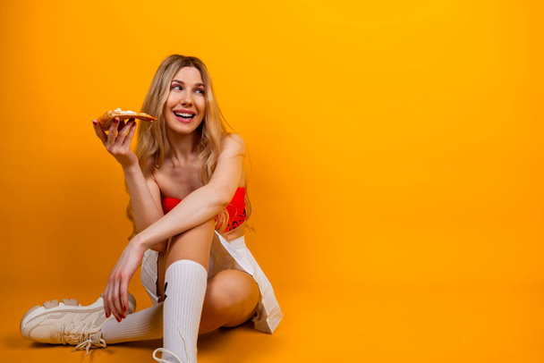 Μια όμορφη αισθησιακή εφαρμογή νεαρή ξανθιά κοπέλα σε ένα κοντό μπλουζάκι και φούστα ποζάρουν με μια φέτα πίτσα. Απομονωμένα σε κίτρινο. - Φωτογραφία, εικόνα