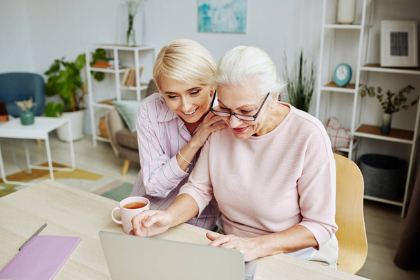 Portrait of smiling young woman helping senior mother using computer in cozy home interior - Photo, Image
