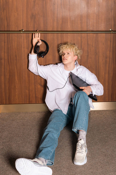 Young trendy teenager, the programmer or IT specialist with a laptop or notebook in hand is sitting in an elevator. Very sad and upset, unhappy. - Photo, image