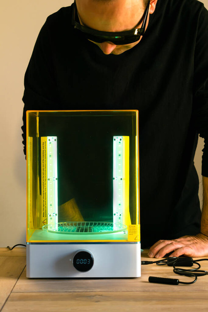 Mies käyttää silmälaseja, jotka säätävät pesu- ja parannuskonetta UV-valon avulla ennen käyttöä 3D-hartsitulostimeen - Valokuva, kuva