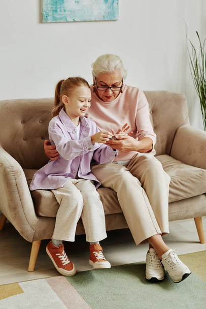 Retrato de larga duración de la niña sonriente mostrando juego móvil a la abuela y enseñándole a usar el teléfono inteligente - Foto, Imagen