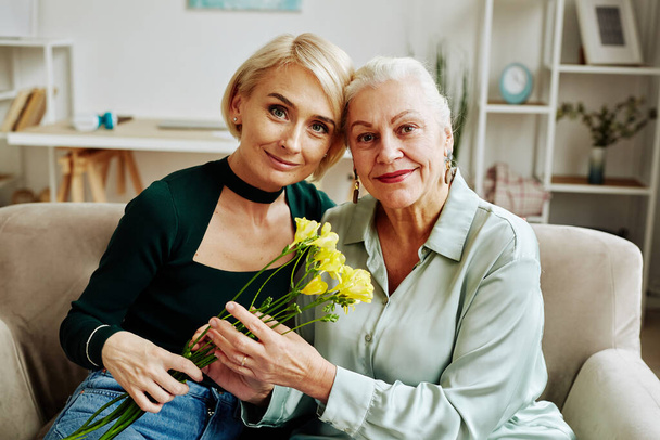 Retrato de vista frontal de hija adulta con madre mirando a la cámara y sosteniendo flores en la acogedora escena casera - Foto, Imagen