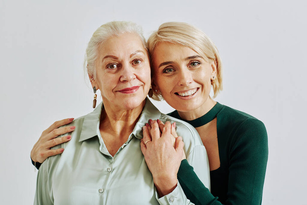 Minimal waist up portrait of smiling adult daughter with senior woman embracing and looking at camera in studio - Foto, afbeelding