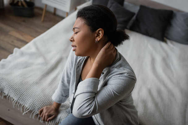 Seitenansicht einer aufgebrachten multirassischen Frau mit Depressionen, die im Sitzen auf dem Bett den Hals berührt  - Foto, Bild