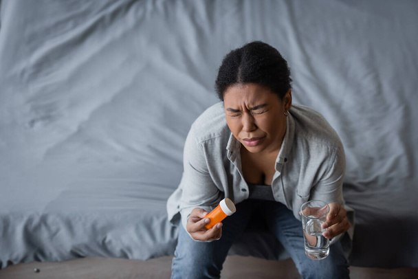 Vue grand angle de la femme multiraciale avec dépression tenant des pilules et de l'eau sur le lit à la maison  - Photo, image