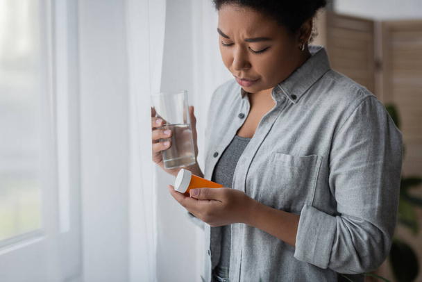 Disappointed multiracial woman with depression holding pills and water near curtain at home  - Foto, afbeelding