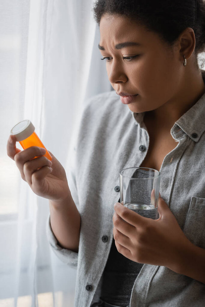 Młoda wielorasowa kobieta z depresją trzymająca tabletki i szklankę wody w domu  - Zdjęcie, obraz