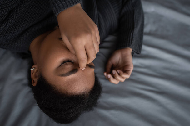 Horní pohled frustrované multiraciální žena dotýkající se obličeje při ležení na posteli  - Fotografie, Obrázek