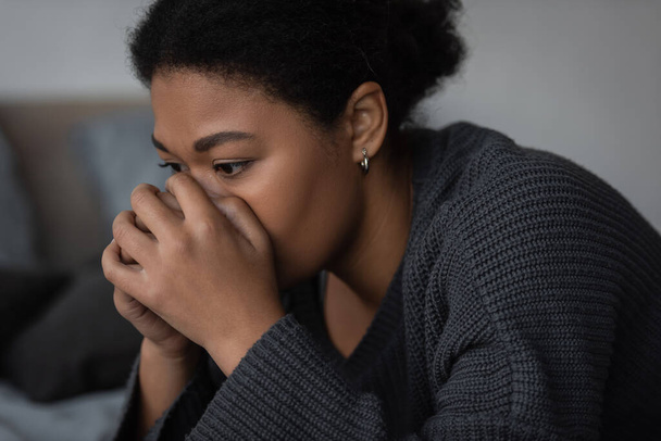 Sad multiracial woman in knitted sweater looking away in bedroom  - Photo, Image