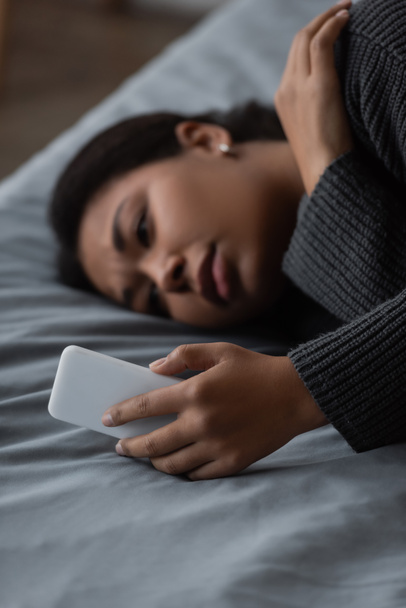 Bulanık, depresyonlu, evde yatarken akıllı telefon kullanan çok ırklı bir kadın.  - Fotoğraf, Görsel