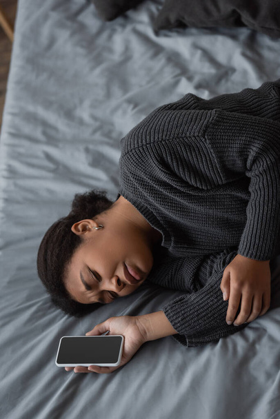 Top view of displeased multiracial woman in sweater holding cellphone on bed  - Photo, image