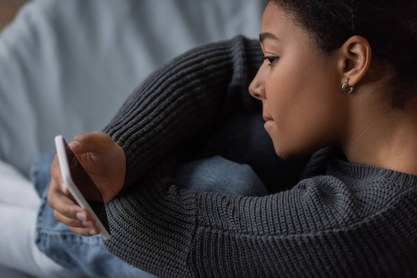 Πλευρική άποψη του κατάθλιψη πολυφυλετική γυναίκα κρατώντας smartphone με λευκή οθόνη στο υπνοδωμάτιο  - Φωτογραφία, εικόνα