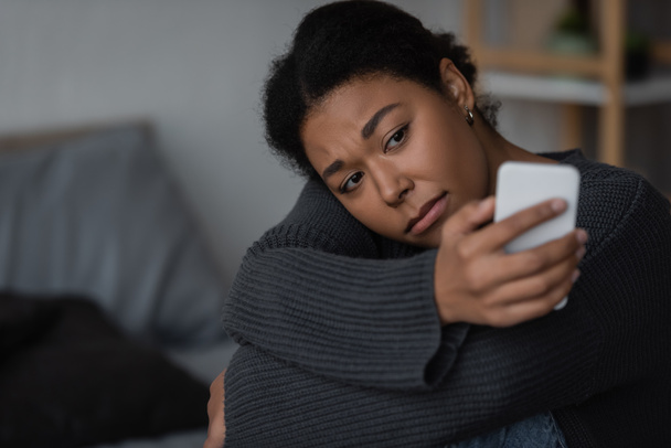 heartbroken multiracial woman in sweater using blurred smartphone in blurred bedroom  - Fotó, kép