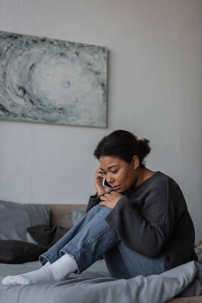Frustrated multiracial woman calling in helpline while sitting on bed at home  - Fotografie, Obrázek