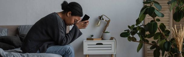 Widok z boku depresji wielorasowy kobieta trzyma telefon komórkowy podczas siedzenia na łóżku w domu, baner  - Zdjęcie, obraz