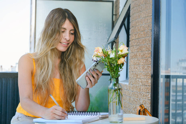 Młoda Latynoska pochodzenia kolumbijskiego uśmiecha się, siedząc na balkonie swojego mieszkania, zapisując w notatniku i korzystając z telefonu, koncepcji biznesowej, przestrzeni do kopiowania. - Zdjęcie, obraz