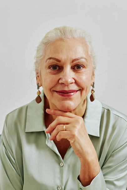 Вертикальный портрет элегантной пожилой женщины, смотрящей в камеру и улыбающейся во время позирования в студии - Фото, изображение
