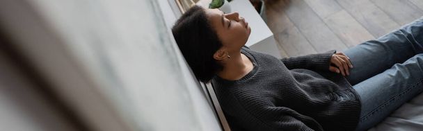 High angle view of depressed multiracial woman leaning on wall while sitting on bed at home, banner  - Fotó, kép