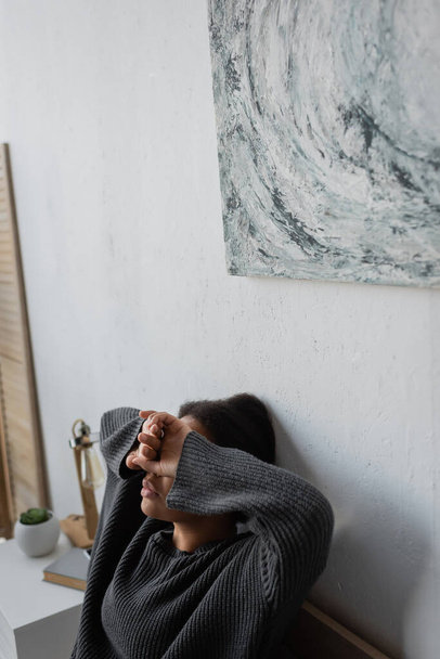 Θλιβερή πολυφυλετική γυναίκα σε πλεκτό πουλόβερ που καλύπτει τα μάτια στην κρεβατοκάμαρα  - Φωτογραφία, εικόνα