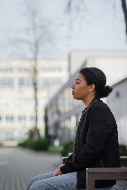 Zijaanzicht van jonge multiraciale vrouw met depressie zittend op bank op stedelijke straat  - Foto, afbeelding