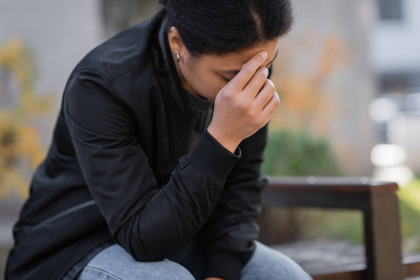 Mulher multirracial deprimida em jaqueta sentado no banco embaçado ao ar livre  - Foto, Imagem