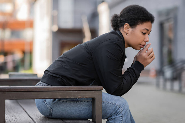 Disappointed multiracial woman in jacket sitting on bench on blurred urban street  - Foto, Bild
