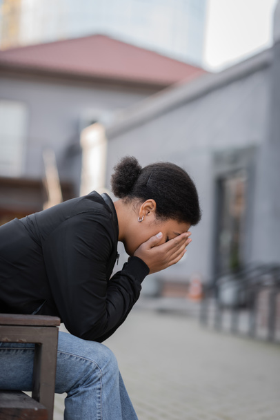 Депрессивная многорасовая женщина покрывает лицо, сидя на скамейке на городской улице  - Фото, изображение
