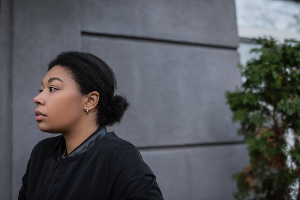 Depressed multiracial woman in jacket looking away near building outdoors  - Foto, imagen