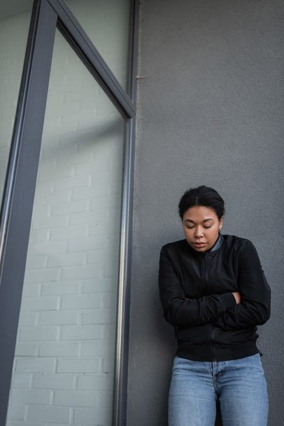 Νεαρή πολυφυλετική γυναίκα με απάθεια στέκεται κοντά σε κτίριο σε εξωτερικούς χώρους  - Φωτογραφία, εικόνα
