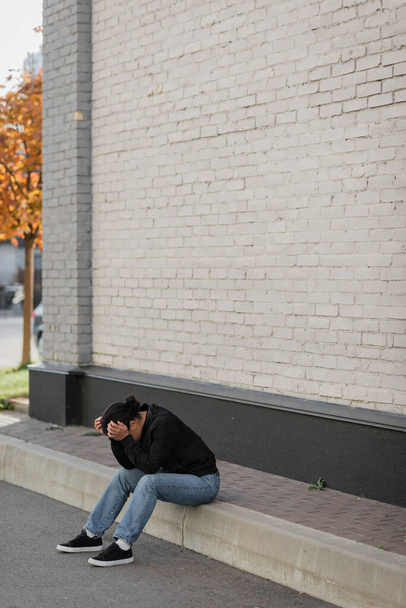 Deprimida mujer multirracial sentada en la frontera cerca del edificio al aire libre  - Foto, imagen