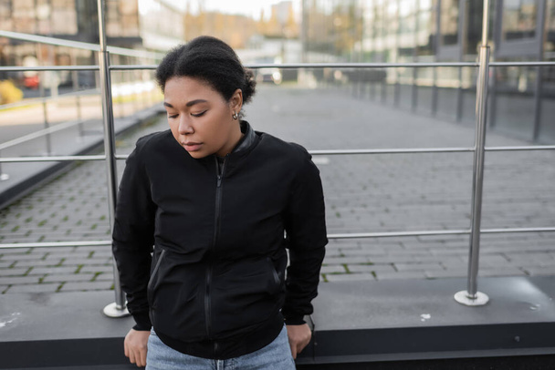 mujer multirracial con problemas mentales de pie cerca de barandilla en la calle urbana  - Foto, Imagen